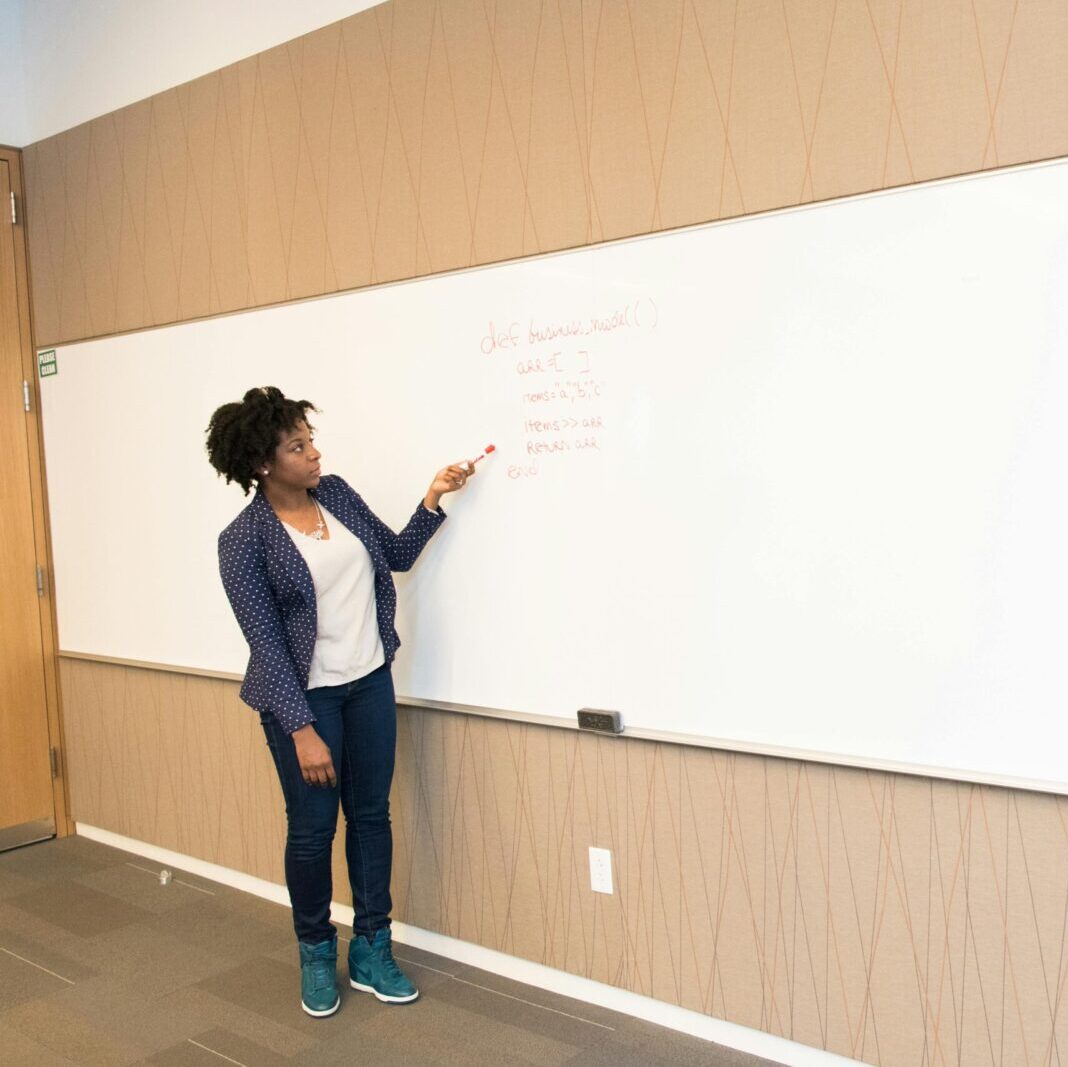 Black female educator explaining concepts on whiteboard in conference room setting.