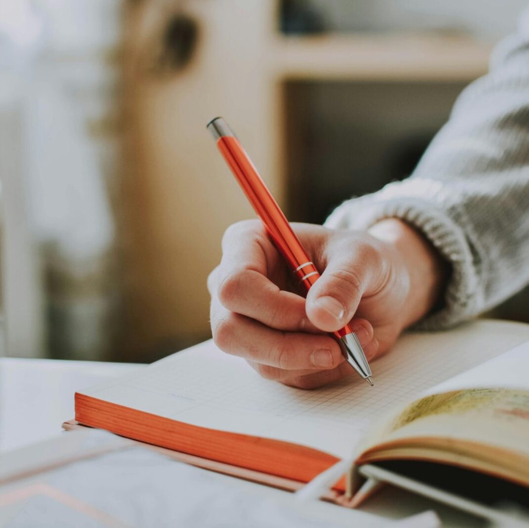 A person writing in a notebook with a red ballpoint pen, showcasing detail and focus.